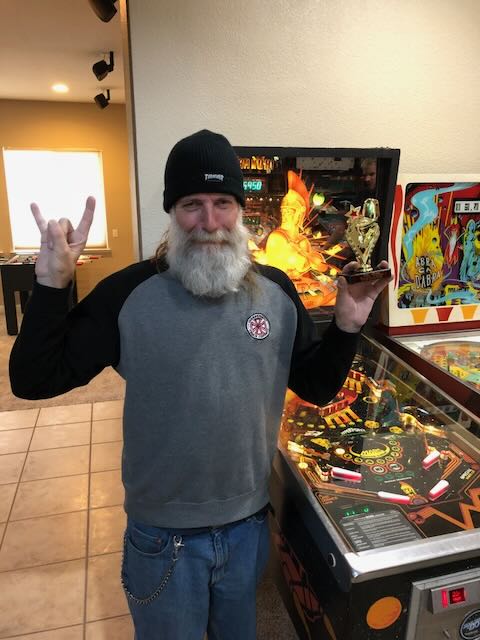 A bearded player in a beanie throwing devil horns beside a lit playfield